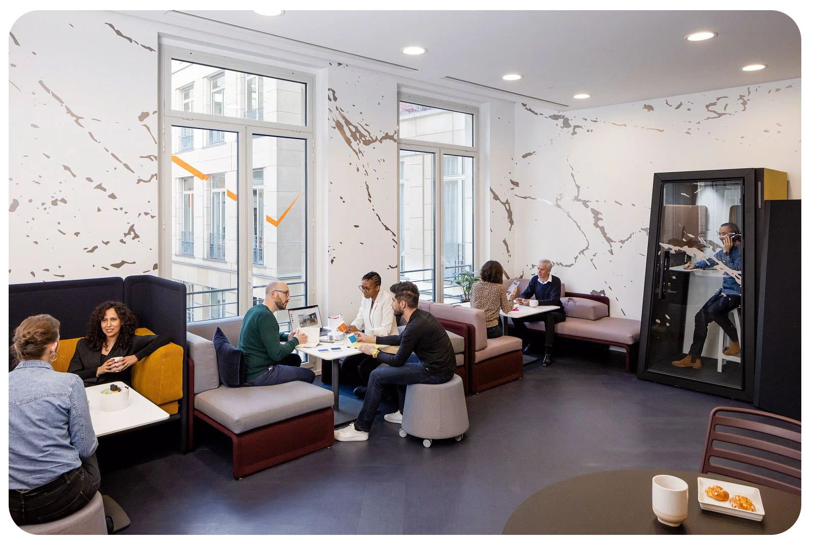 interior lounge area of a workplace. people are seating in groups of 2-3, collaborating or discussing. Next to the far wall there is an acoustic booth and someone is inside talking on the phone.